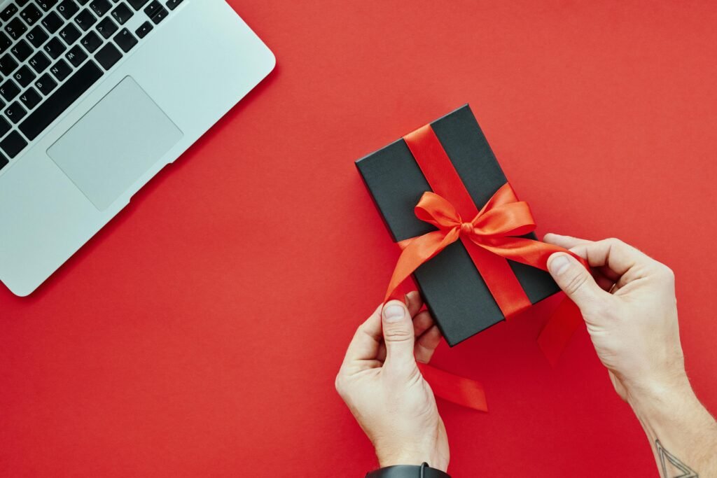 Holiday Tech Deals (2025): The Easy Shortlist Close-up of hands tying a red ribbon on a gift box beside a laptop on a red background, perfect for holiday presents.